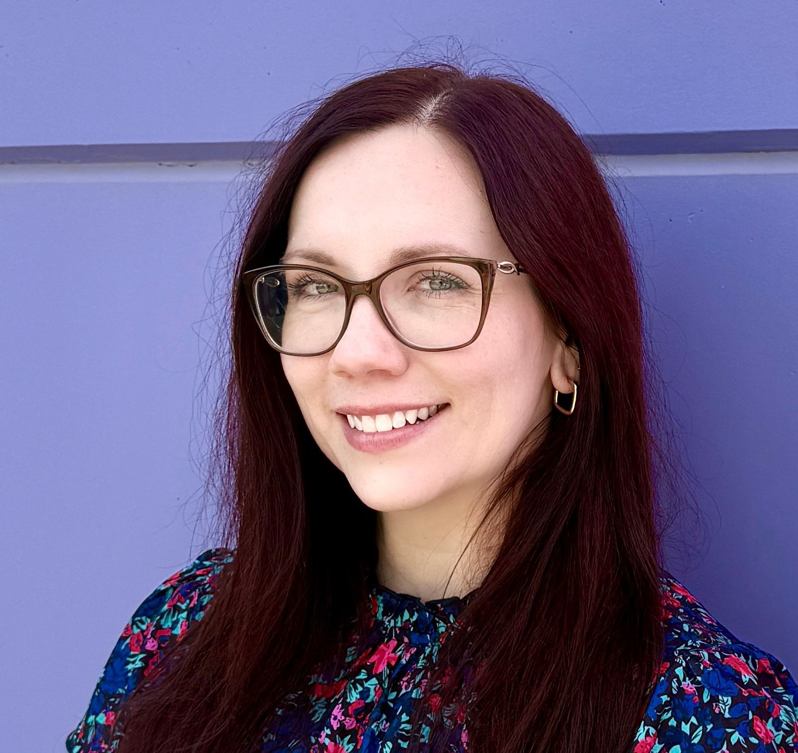 Woman with glasses smiles in front of a purple background, wearing a floral top.