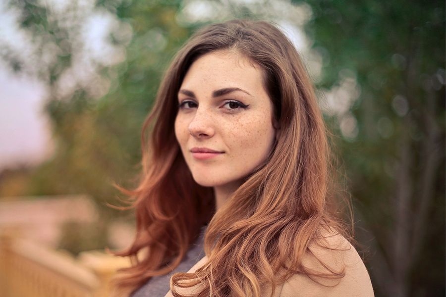 Woman with auburn hair, freckles, and a slight smile, outdoors with a blurred green and tan background.