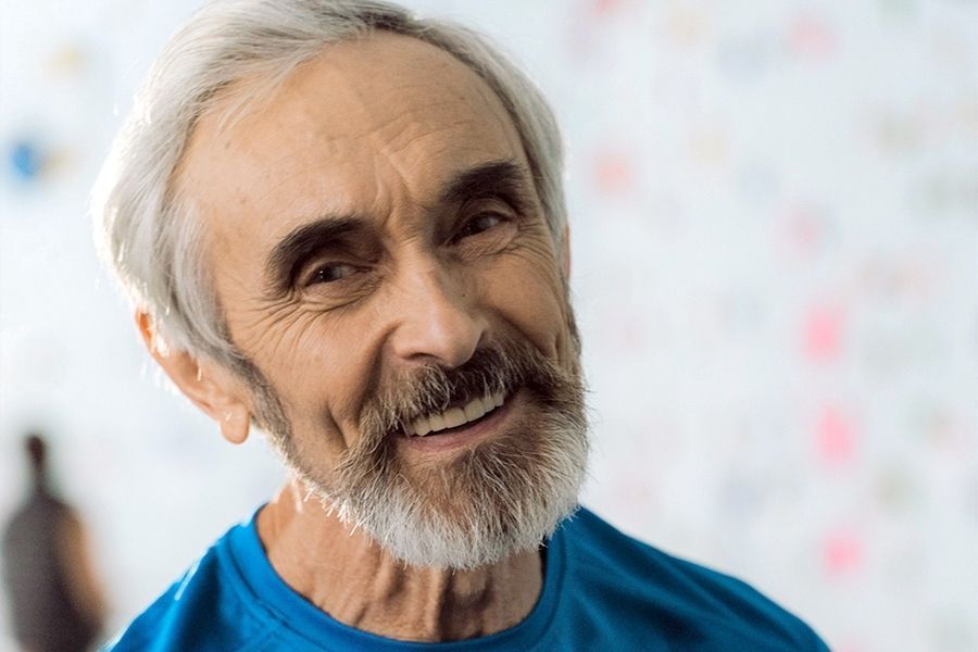 Smiling older man with gray hair and beard, wearing a blue shirt, against a blurred white background.