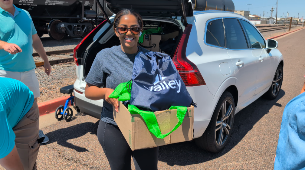 Group of volunteers packing boxes outdoors, smiling, wearing green shirts, sunny park setting.