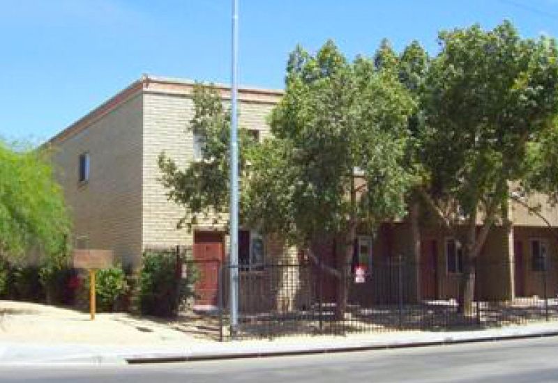 Two-story beige building with trees in front; sidewalk and street below, blue sky overhead.