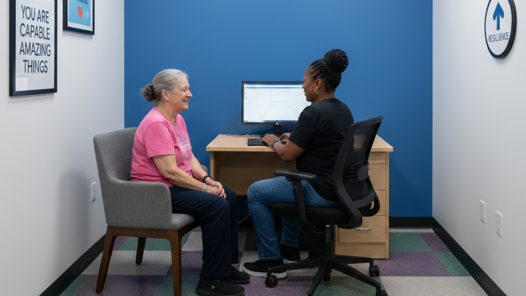 A woman in pink shirt smiles, talking with another at desk. Blue wall, computer, framed art, office setting.