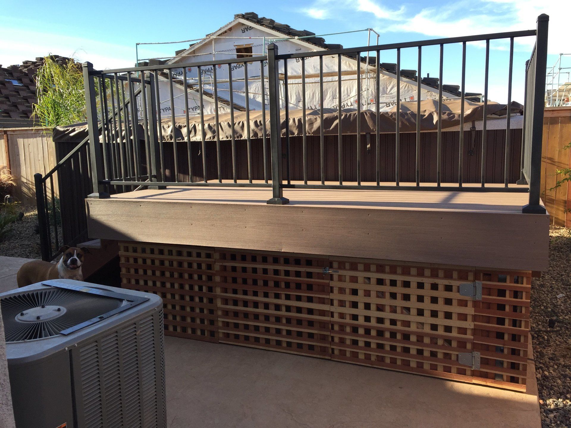 A dog is sitting on a deck next to an air conditioner.