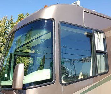 Close-up of a brown RV exterior, showing the windshield, side window, and side mirror against a sunny background.