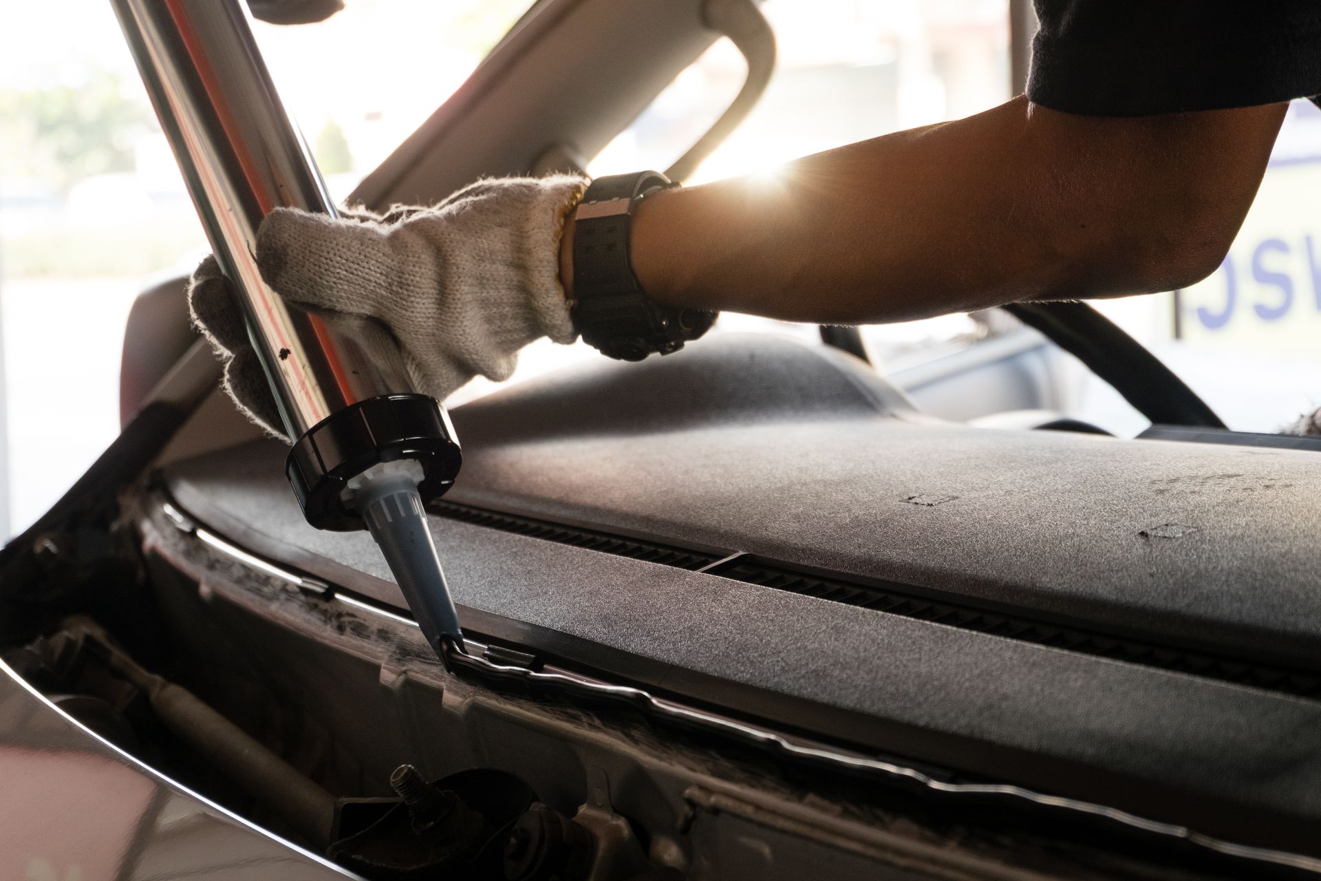 A person in work gloves is applying sealant to a car's windshield frame with a caulking gun.