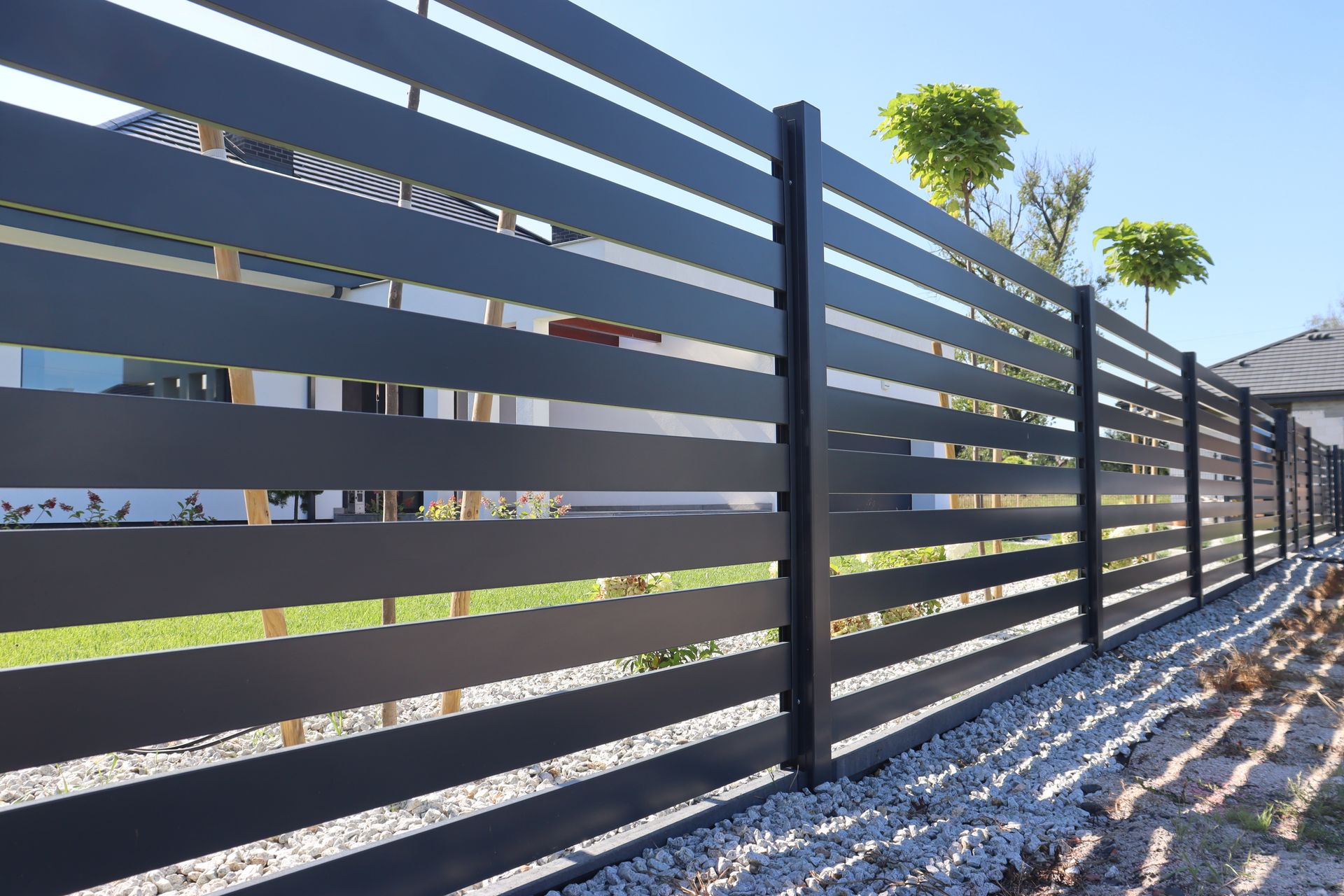 A modern black horizontal slat fence lines a residential property with a green lawn and gravel ground.