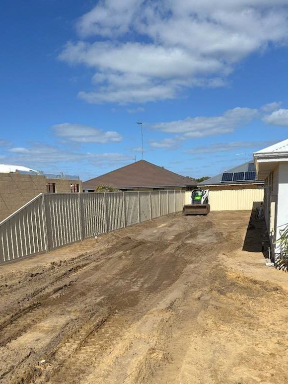 A small roller compacts dirt in a backyard, next to a fence and house, under a blue sky.