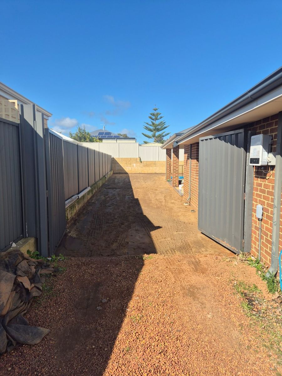 Narrow gravel path between gray fences and brick building under a blue sky.