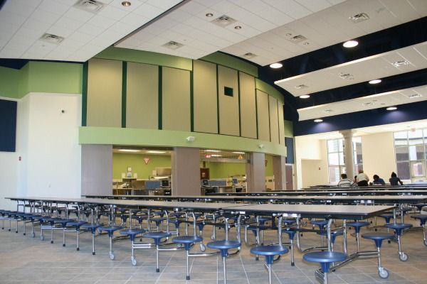 School cafeteria with long tables, blue chairs, and food service area. Light green and blue walls.