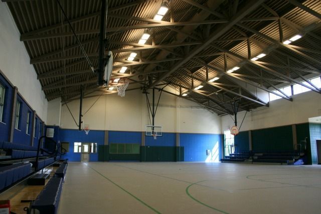 Indoor gymnasium with basketball hoops, blue and green walls, wooden ceiling, and bleachers.
