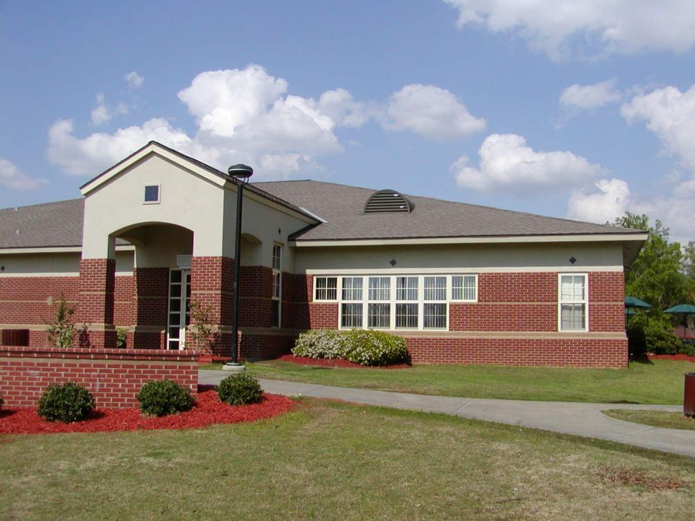 Brick building with a beige entrance, windows, and a dark roof, set on a grassy lawn under a blue sky.