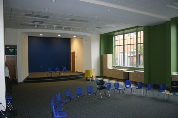 A blue stage with chairs arranged in a circle, a large window, and a green wall.