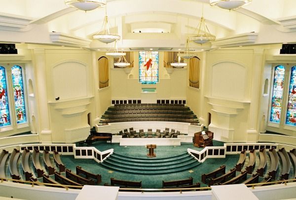 Church interior with rows of pews facing a stage with a pulpit and organ. Stained glass windows and teal carpet.