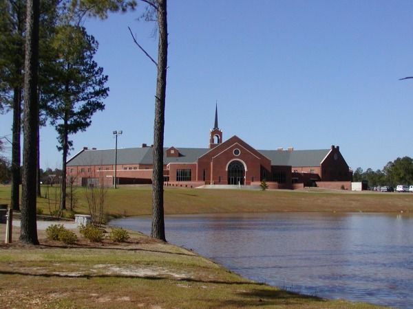 Red brick church building with a steeple, reflecting in a pond, under a blue sky.