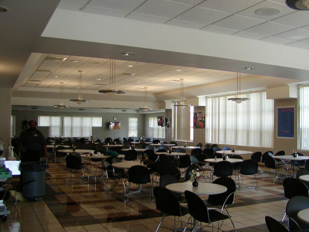 Cafeteria with many round tables, chairs, and large windows. A person stands near a trash can.