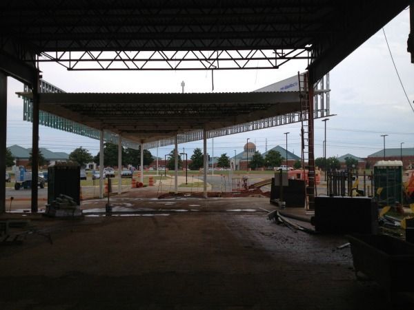 Construction site with open roof structure, exterior view; cloudy sky, distant buildings.
