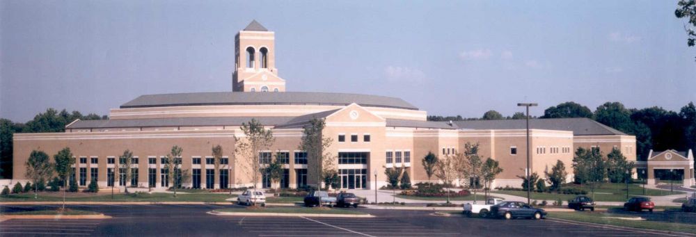 Beige building with a tall tower and arched entrance. Parking lot in the foreground, trees in the background.