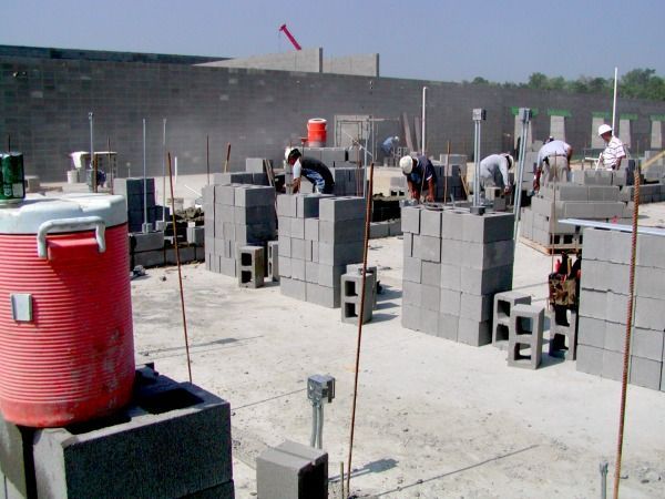 Construction workers laying concrete blocks outdoors; sunlit, red cooler in foreground.