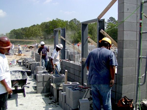 Construction workers building a cinder block wall outdoors. Several men in hard hats, laying bricks, and using tools. Sunny day.