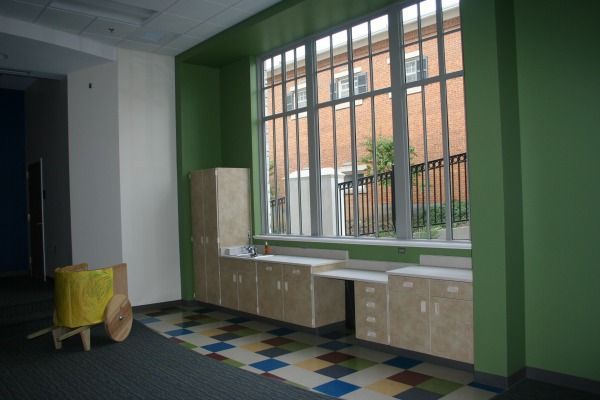 A bright green-walled room with cabinets under a large window. A toy wagon sits nearby on a colorful tiled floor.