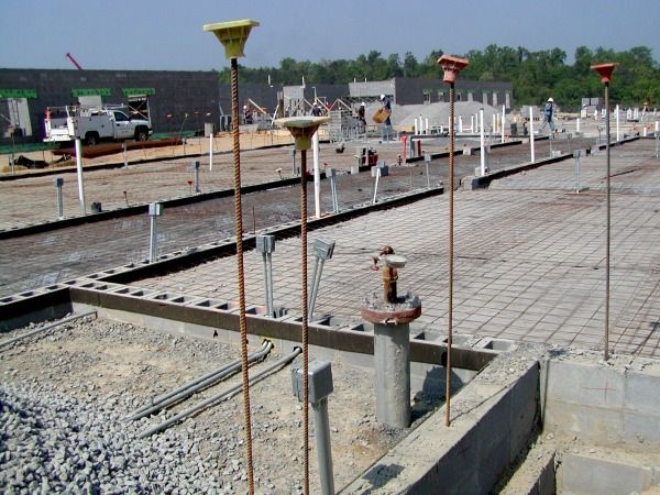 Construction site with concrete forms, rebar, and utility pipes; orange and yellow markers stand upright.