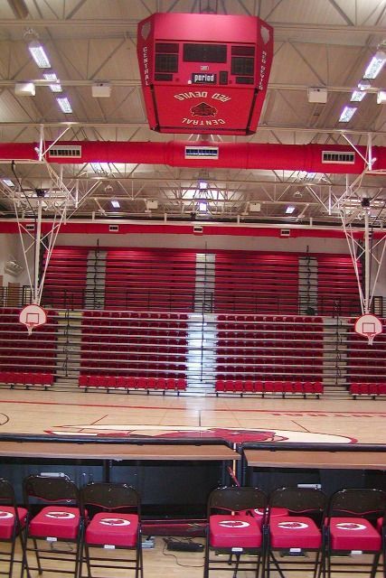 Interior of a gymnasium with red bleachers, scoreboard, and basketball court. Folding chairs in foreground.