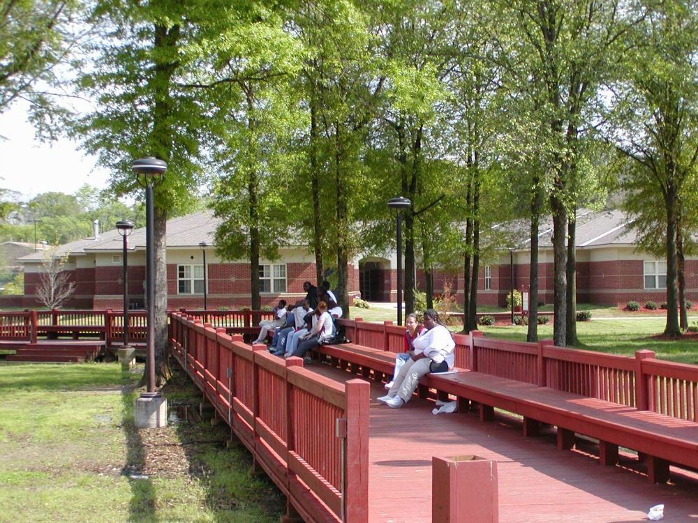 People sitting on red benches, near a brick building with trees in the background.