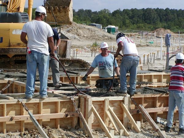 Construction workers pouring concrete into forms on a construction site; excavator in background.