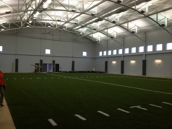 An indoor football practice field with green turf, white lines, and a high, white ceiling.