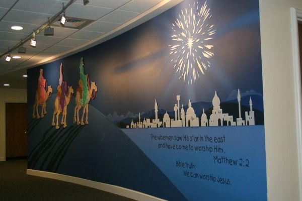 Mural of the three wise men on camels, fireworks, and a city skyline with a bible verse on a blue wall.