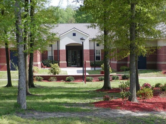 A one-story building with red brick and a tan facade, framed by trees on a grassy lawn.