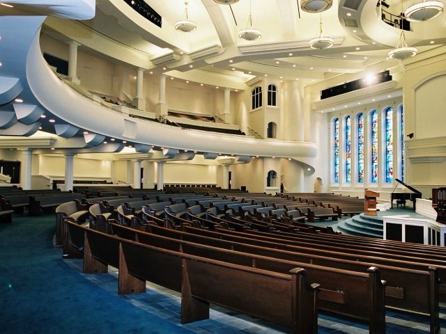 Interior of a large church with rows of pews, blue carpet, and a stage with a piano and stained glass windows.
