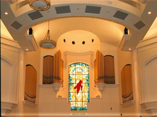 Interior of a church. Organ pipes flank a stained glass window with a red bird. Soft lighting and beige walls.