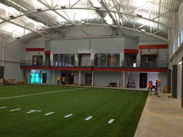 Indoor football field under construction, featuring artificial turf, red accents, and workers.