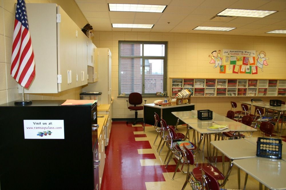 Classroom with desks, cabinets, American flag, and teacher's desk. Red, white, and beige color scheme.