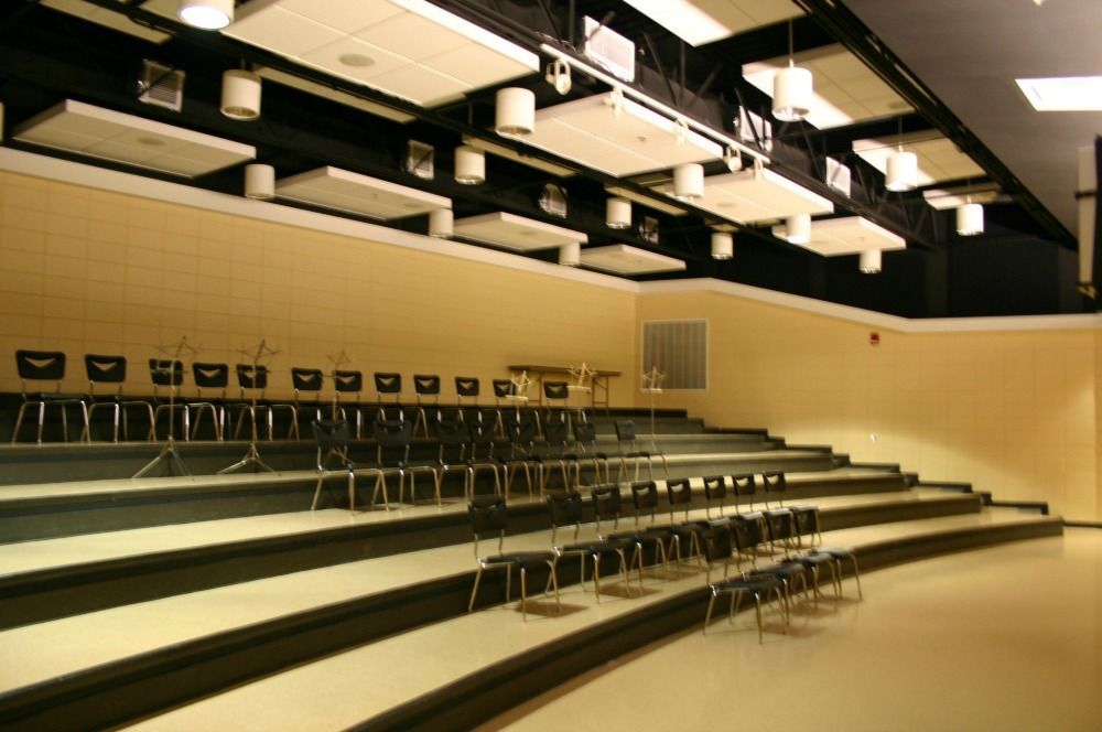 Auditorium with tiered seating, black chairs, beige walls, and acoustic panels on the ceiling.