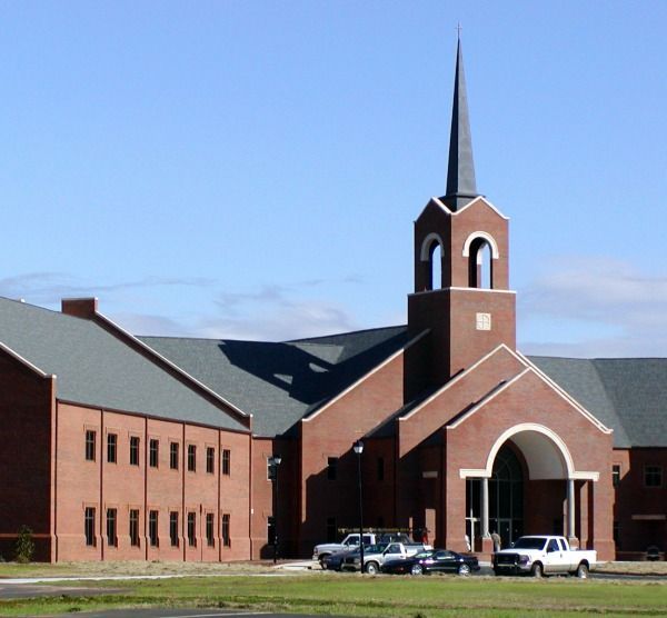 Red brick church with a steeple under a blue sky. Vehicles are parked in front.