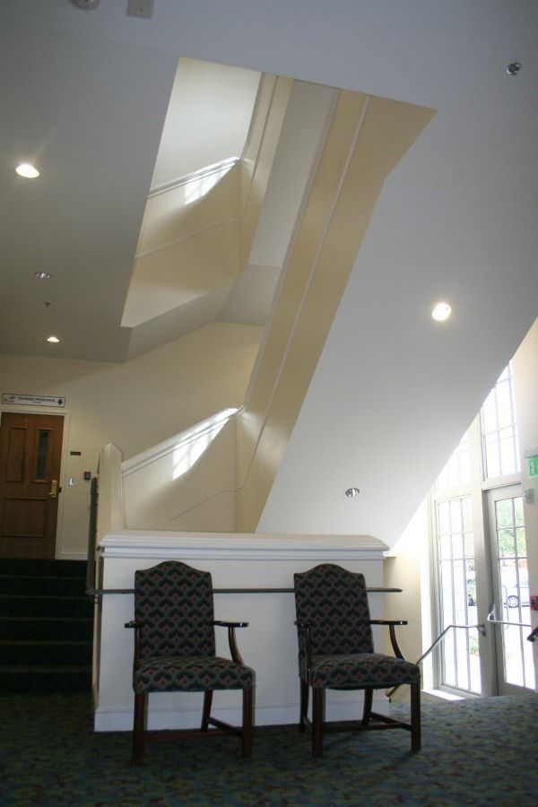 Two chairs in a hallway below a winding staircase, bathed in light. White walls, dark carpet.