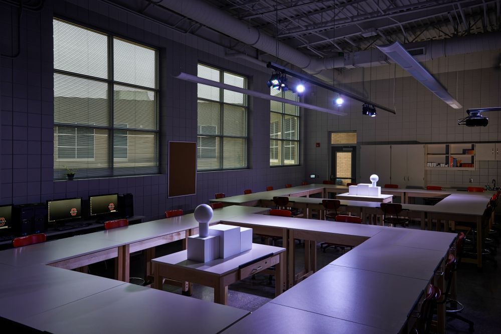 An art classroom with tables arranged in a U-shape. Windows let in light, illuminating sculptures and computers.