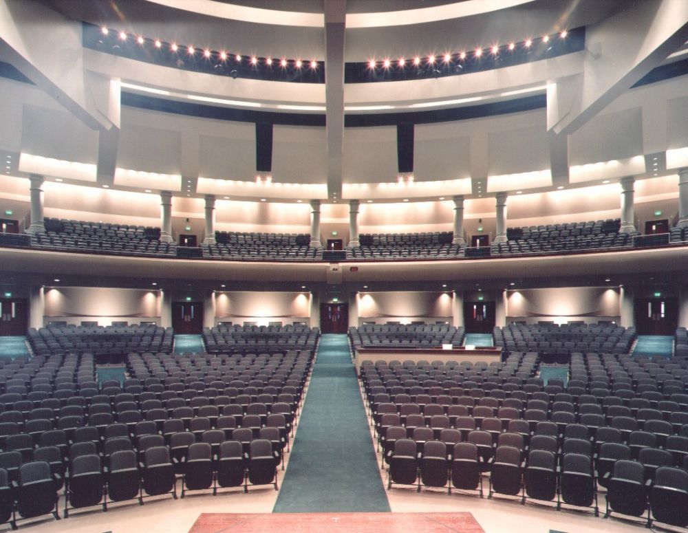 Auditorium interior. Rows of seats face a stage. Beige walls, blue carpet.