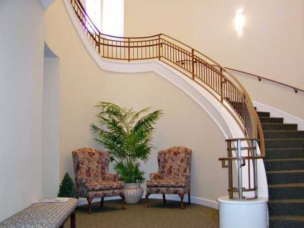 Two patterned armchairs and a potted plant sit beneath a curved staircase with brown railing.