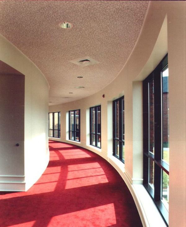 Curved hallway with red carpet, windows, and white walls. Ceiling has recessed lighting.