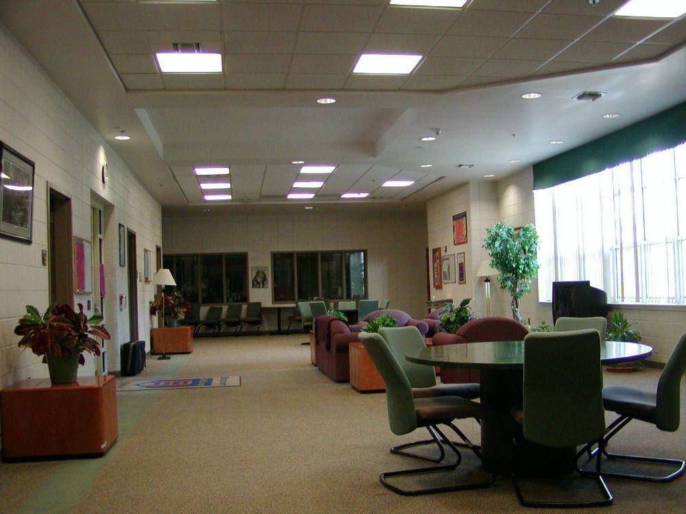 Lounge area with seating, tables, and plants. Light brown carpet, off-white walls and a large window.