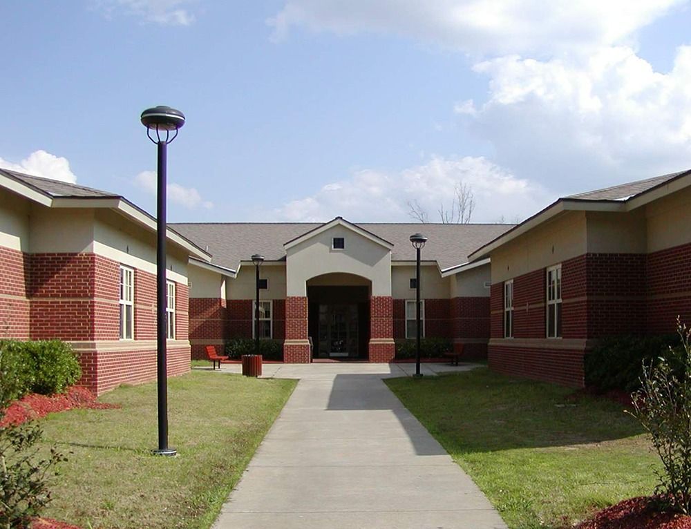 Exterior view of a brick building with a walkway leading to a dark doorway, grassy lawn, and blue sky.