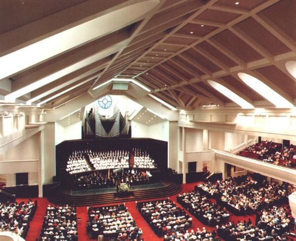 A church interior with a choir on stage, rows of seated people, and a high, angled ceiling.