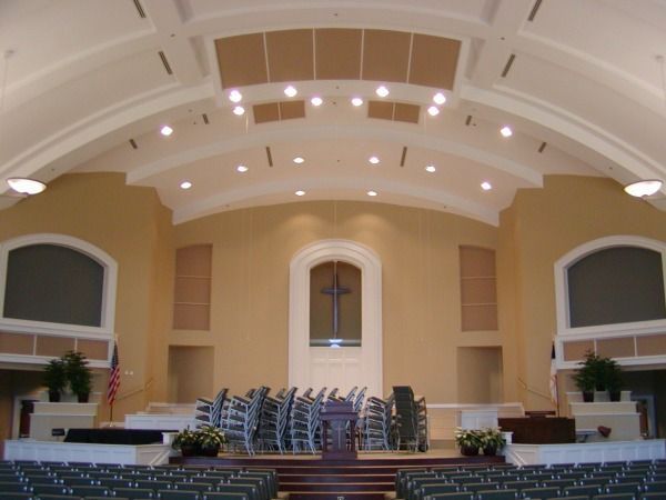 Interior of a church with tan walls, a stage, and chairs. A cross hangs above the stage.