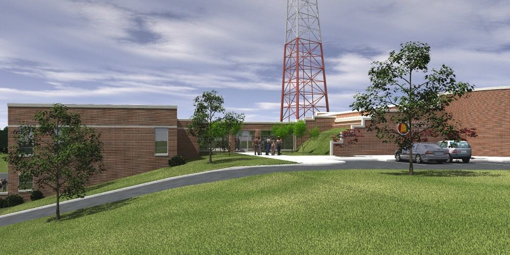 A red brick building with a tall radio tower in the background, cars parked near the entrance, and a grassy hill in front.