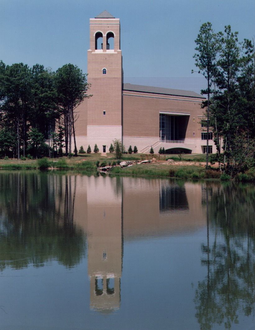 Beige church tower reflected in a still pond. Blue sky, trees, and shrubbery.