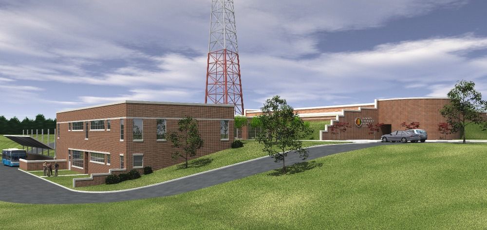 Brick buildings and a radio tower on a grassy hill under a cloudy sky; a vehicle drives on a road.