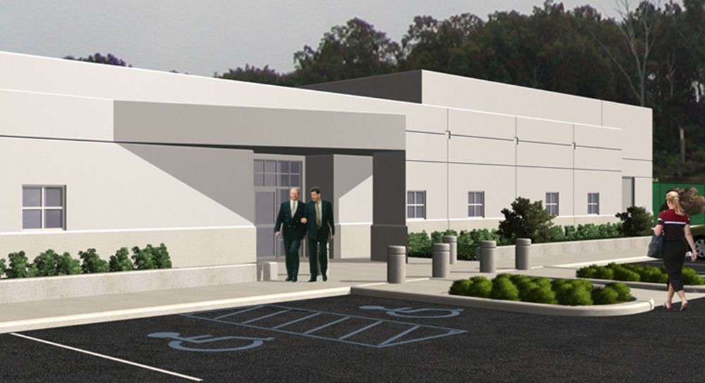 Exterior view of a building with people entering. Gray building, accessible parking, bushes, and trees.